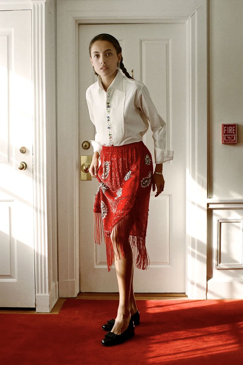 Model in a white buttoned shirt and beaded red fringed skirt standing in a hotel hallway with red carpet