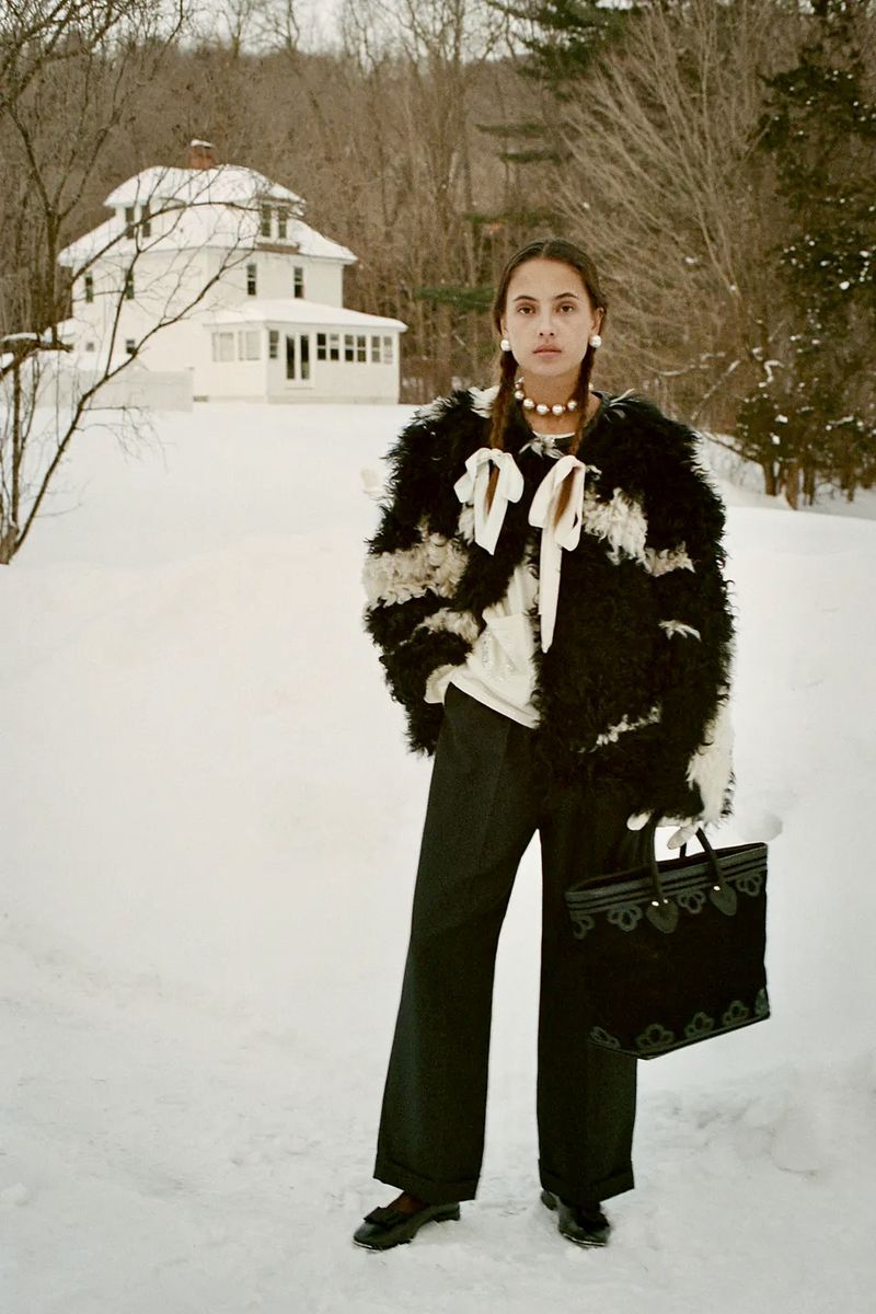 Model in a black textured jacket holding a tote bag standing outdoors in a snow-covered landscape with a white house in the background.