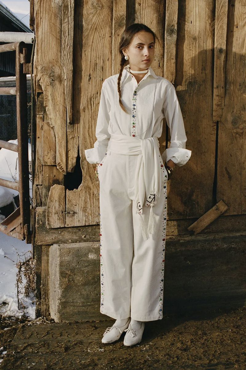 Model in white long-sleeve button-down shirt and matching trousers standing against a weathered wooden barn exterior