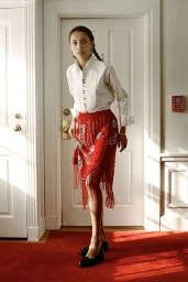 Model in a white buttoned shirt and beaded red fringed skirt standing in a hotel hallway with red carpet