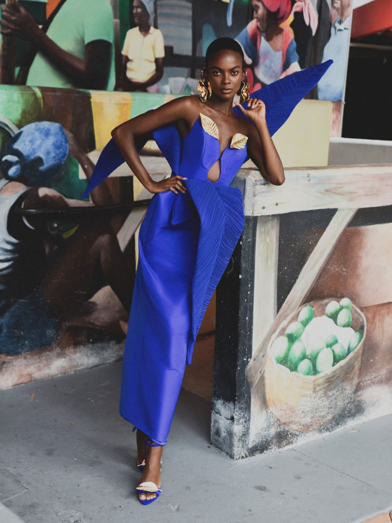 Black woman modeling a structured blue avant-garde dress with sculptural wing-like shoulders against a mural background