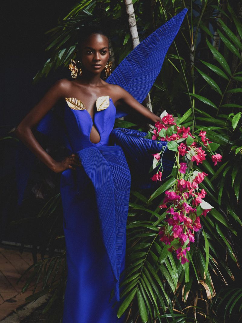 Model Aube Jolicoeur in a cobalt blue sculptural leaf gown with gold accents posing among tropical palm foliage.