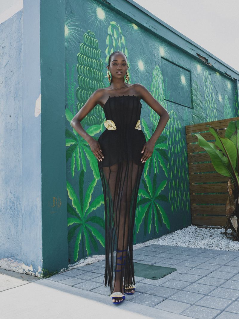 Model in a black strapless sheer dress posing against a teal mural with green painted palm tree patterns.