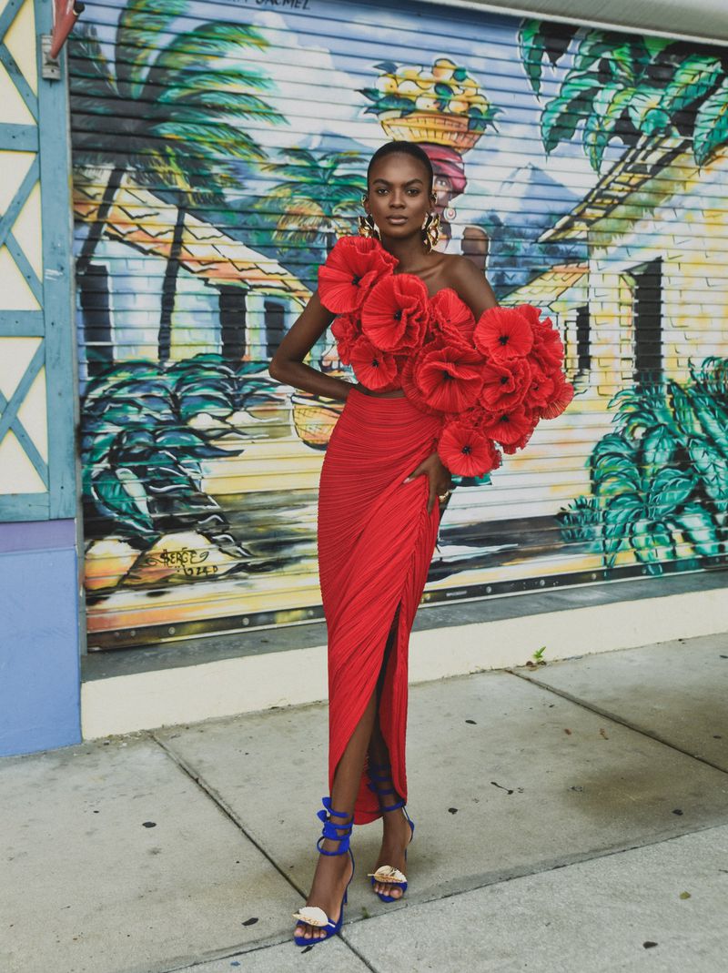 Model Aube Jolicoeur posing in a sculptural red floral rosette dress with blue strappy heels against a painted street mural.