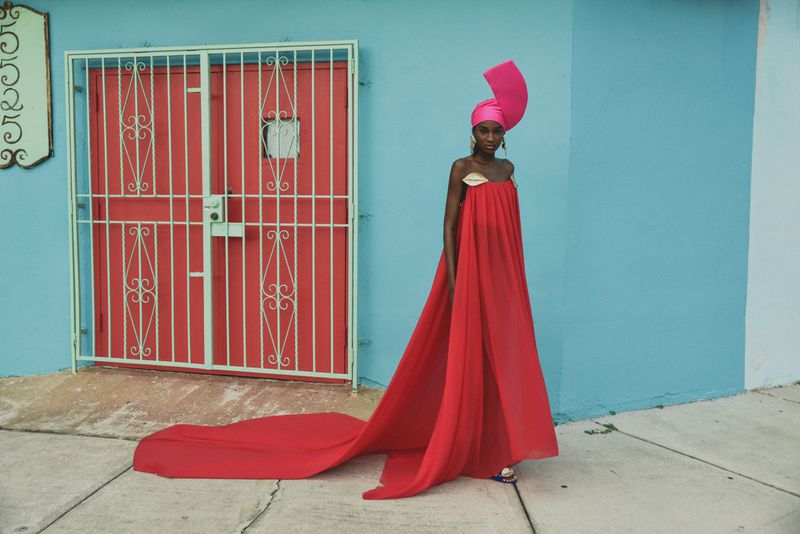 Model wearing a flowing red strapless gown with a long train and a sculptural pink headpiece against a turquoise wall and red gate.