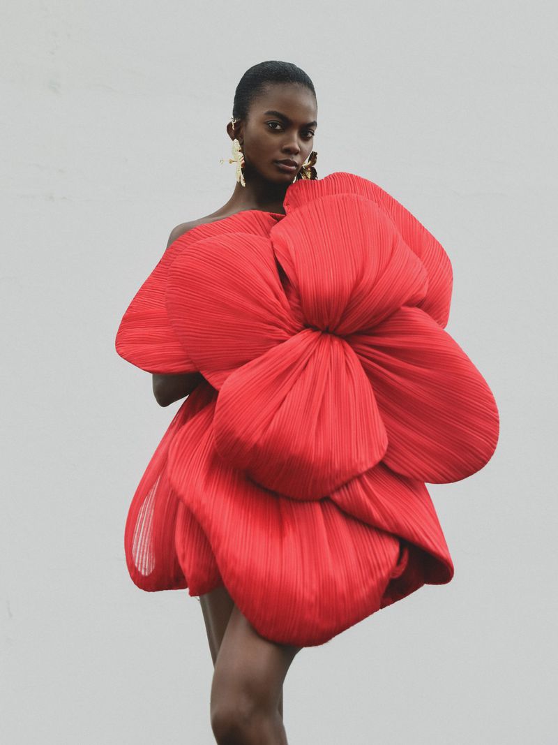 Model wearing a sculptural red pleated flower dress with large statement earrings against a neutral grey studio background