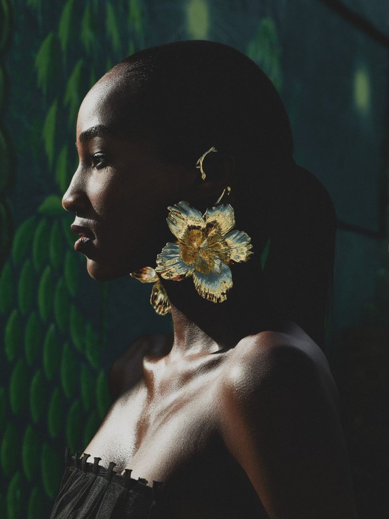 Profile portrait of a black woman wearing oversized gold floral earrings and a black strapless top against a dark green backdrop.