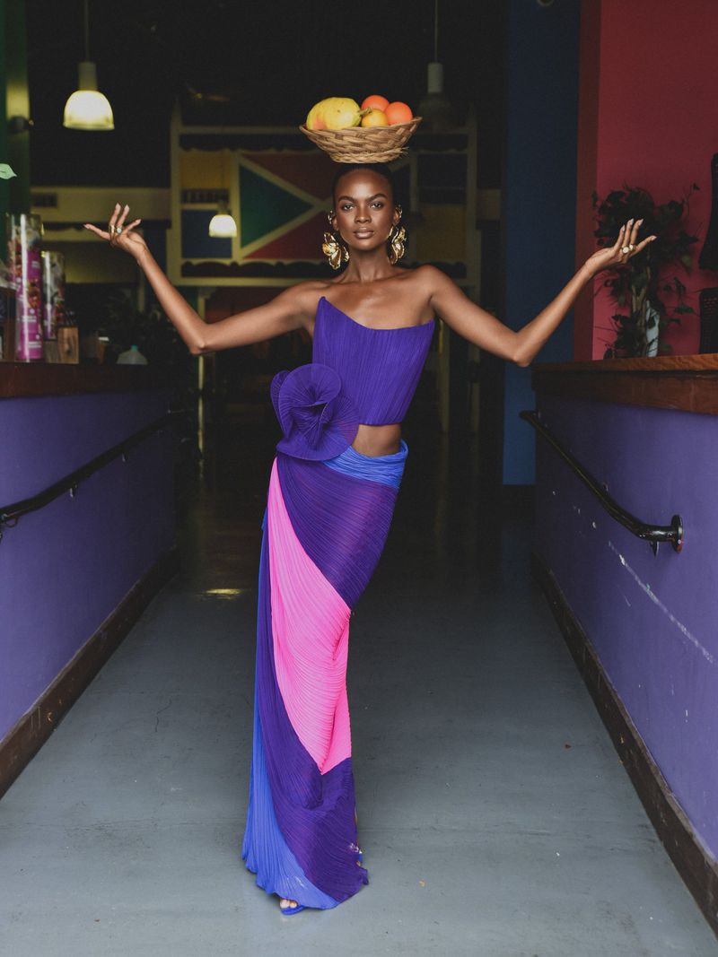 Model in a purple pleated dress balancing a basket of fruit on her head in an indoor hallway setting