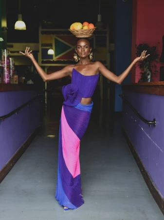 Model in a purple pleated dress balancing a basket of fruit on her head in an indoor hallway setting