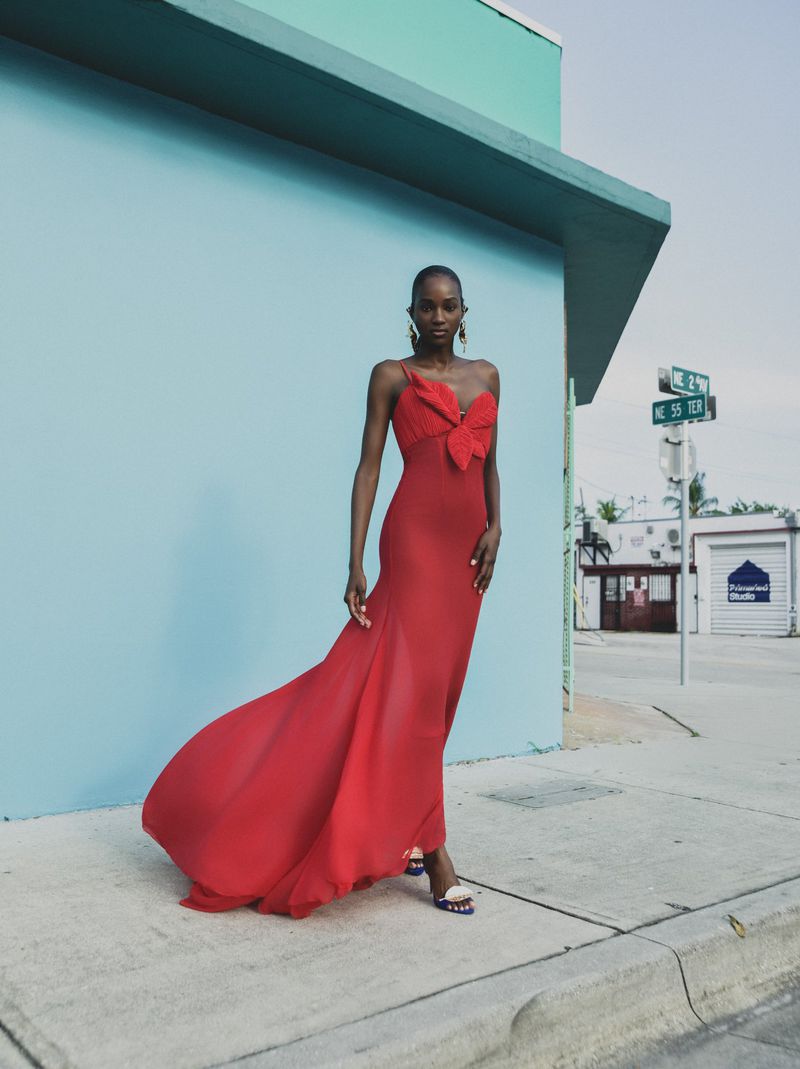 Model in a long red dress standing outdoors on a sidewalk against a bright blue wall for Atelier Ndigo spring summer 2026
