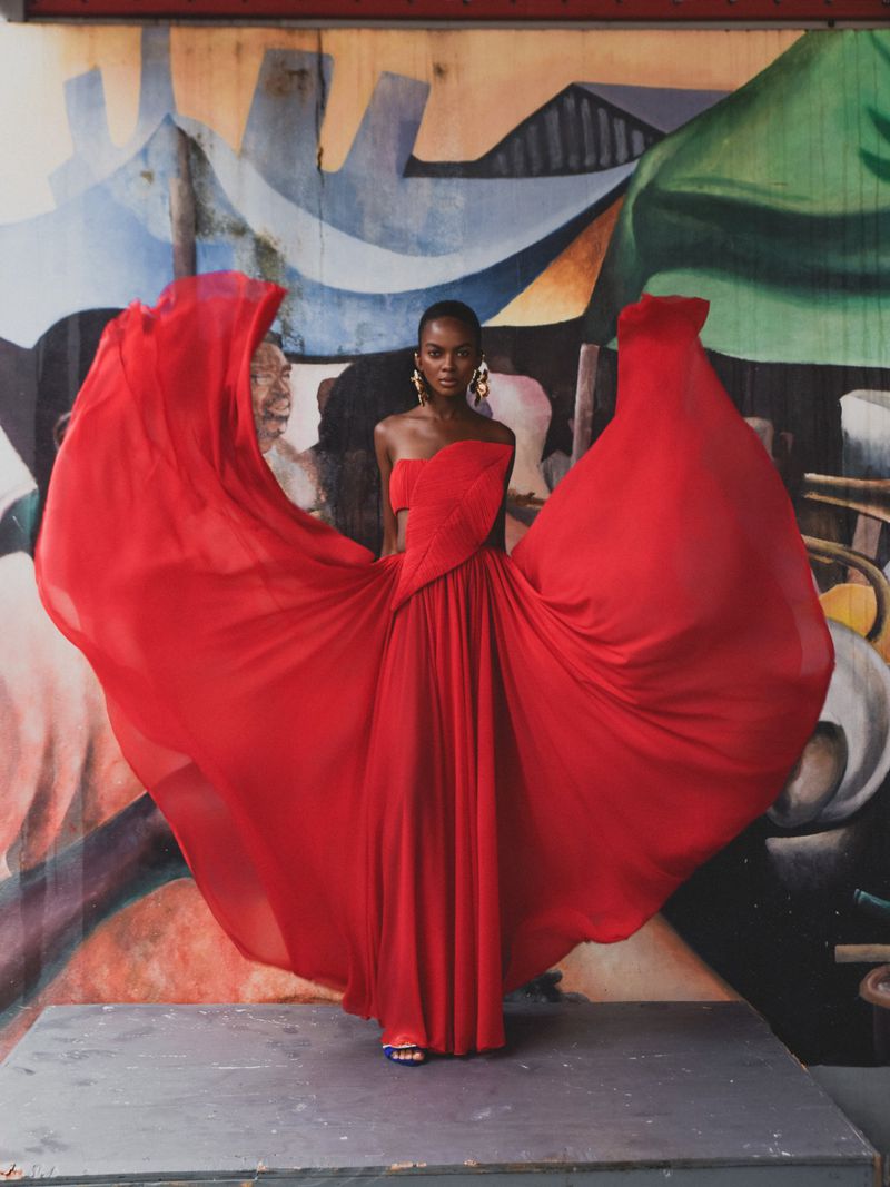 Model in a flowing strapless red gown with expansive draped fabric standing before a colorful painted mural.