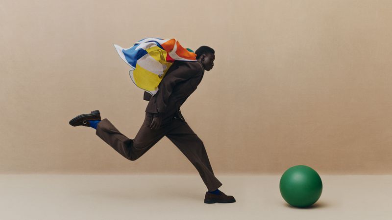 Model leaping in a dark brown suit with a colorful printed scarf billowing behind him near a green sphere against a beige backdrop.