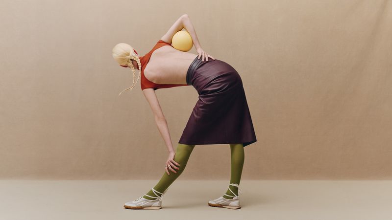 Model in a backless top and burgundy leather skirt bending forward with a yellow ball against a beige backdrop.