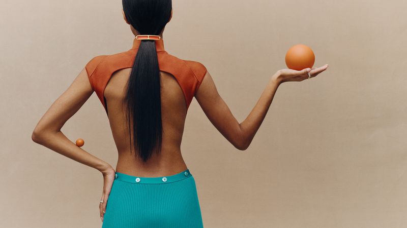Model with long dark hair wearing a backless rust-orange top and turquoise skirt holding an orange sphere