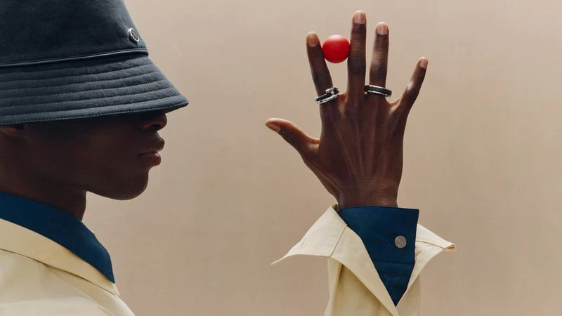 A Black model in a dark bucket hat and pale yellow jacket raises his hand, balancing a red ball between fingers adorned with silver rings.
