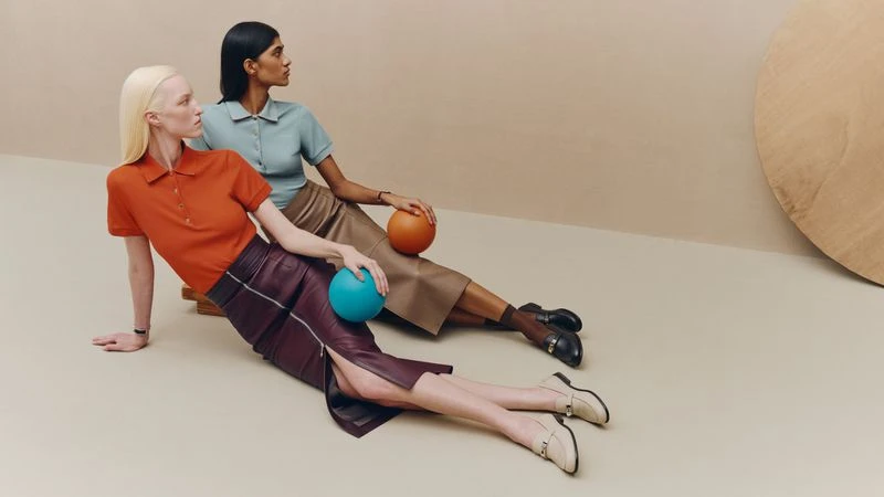 Two female models posing seated on a neutral studio floor holding small colored spheres, wearing Hermes fashion styling.