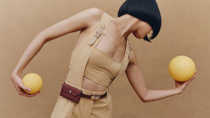 Model with a black bob posing in a beige buckled outfit while holding two yellow spheres against a warm neutral backdrop.