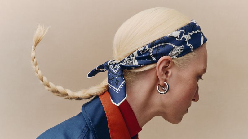 Model with platinum blonde hair and a braided ponytail wearing a blue printed silk scarf and hoop earrings