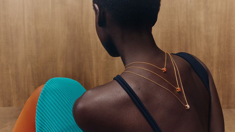 Woman showcases gold necklaces with orange stone details, wearing a black top and brightly colored bottoms against a wood backdrop.