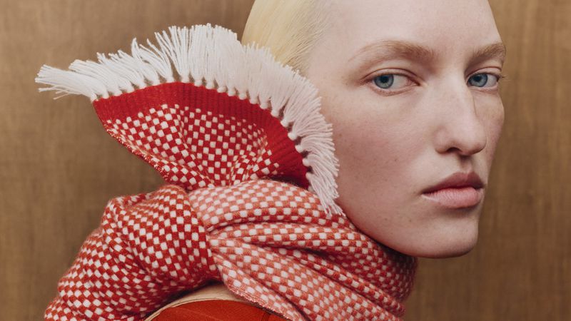 Close-up portrait of a blonde model wearing a red and white patterned fringed scarf for Hermes Edito SS26.