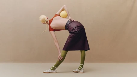 Model in a backless top and burgundy leather skirt bending forward with a yellow ball against a beige backdrop.