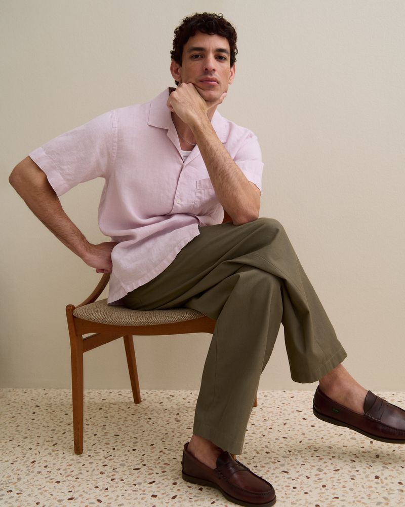 Man seated on a wooden chair wearing a pale pink short-sleeve shirt, olive trousers, and brown loafers over a terrazzo floor.