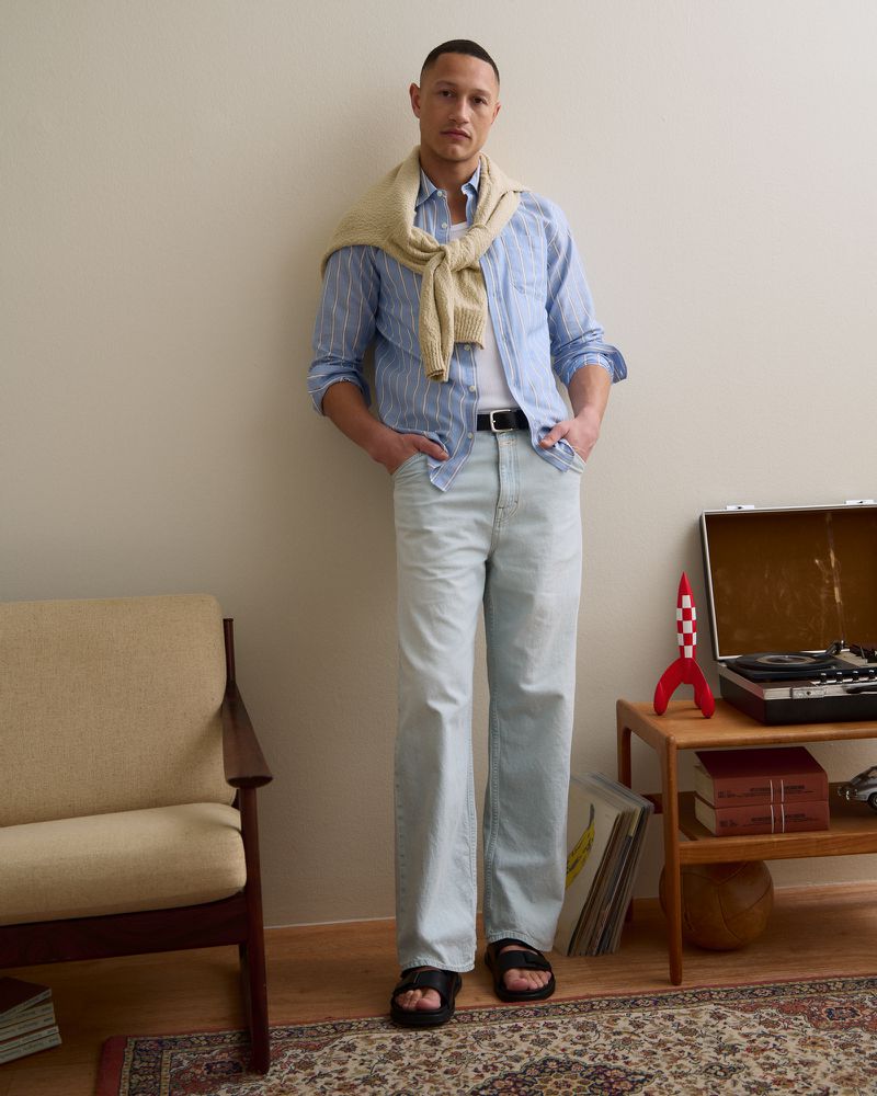 Model wearing a blue striped shirt, light wash denim jeans, and a cream sweater over shoulders in a living room setting.