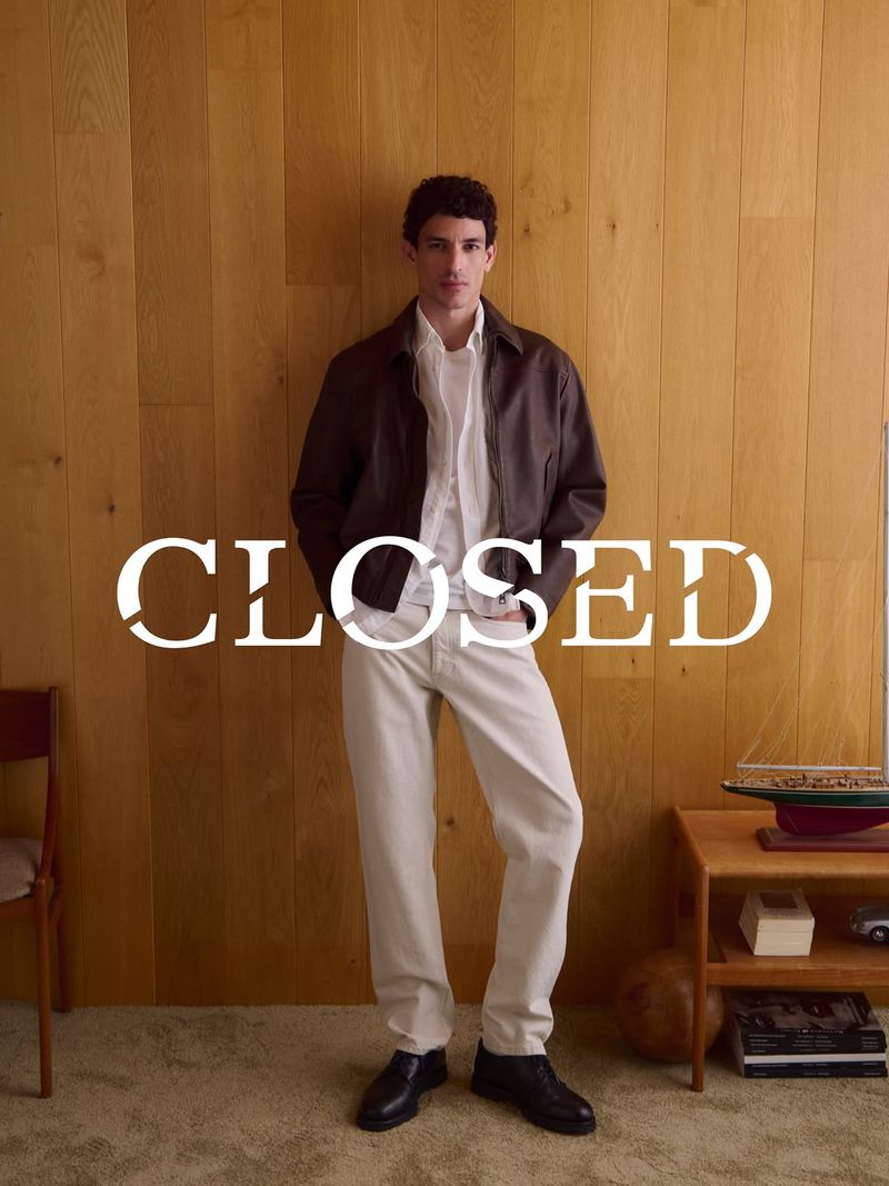 Model standing against a wood panel wall wearing a dark jacket, white shirt, and light trousers for Closed.