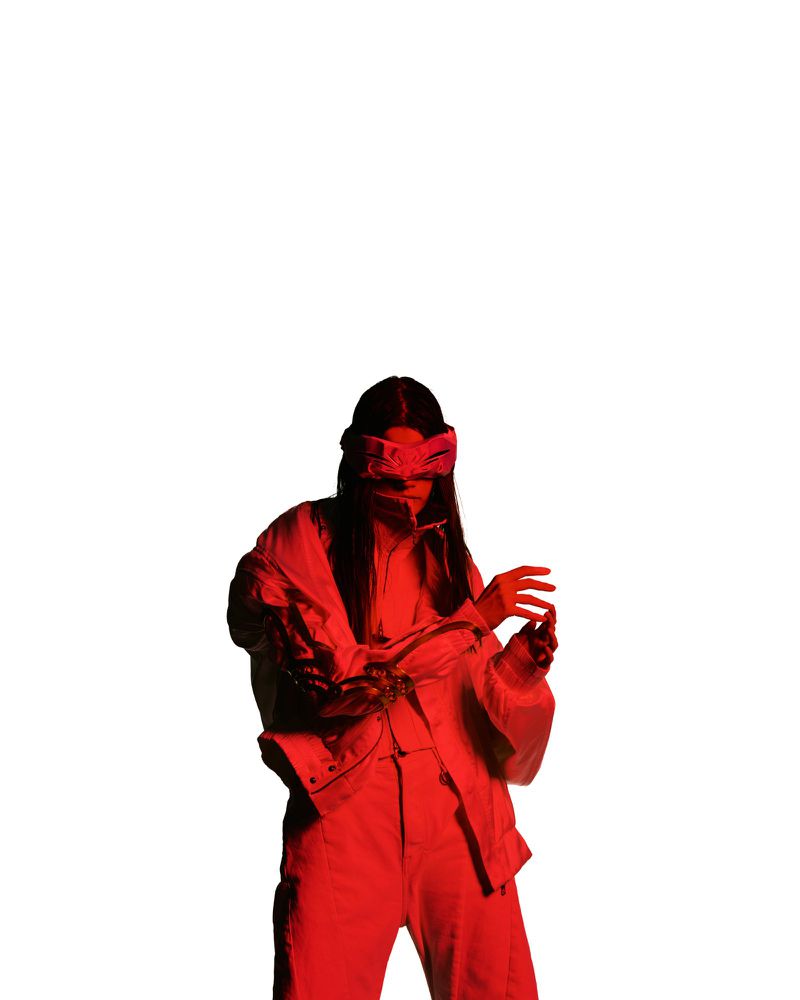 A model wearing layered clothing and a head accessory illuminated by intense red lighting against a clean white background