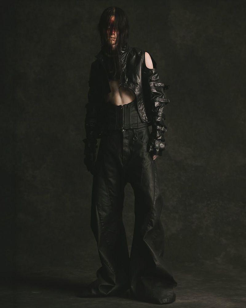 Model in an avant-garde cropped black jacket, corset belt, and wide-leg trousers posing against a dark textured backdrop.