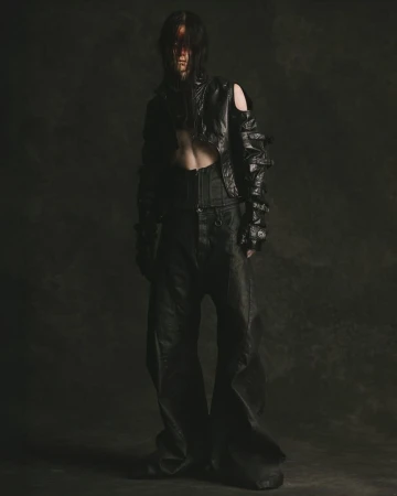 Model in an avant-garde cropped black jacket, corset belt, and wide-leg trousers posing against a dark textured backdrop.