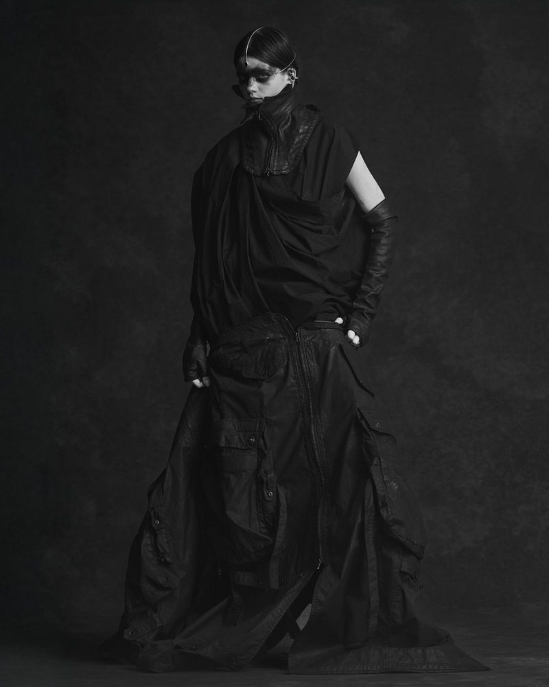 Model wearing a draped all-black avant-garde outfit with long gloves and dark face paint against a textured studio backdrop.