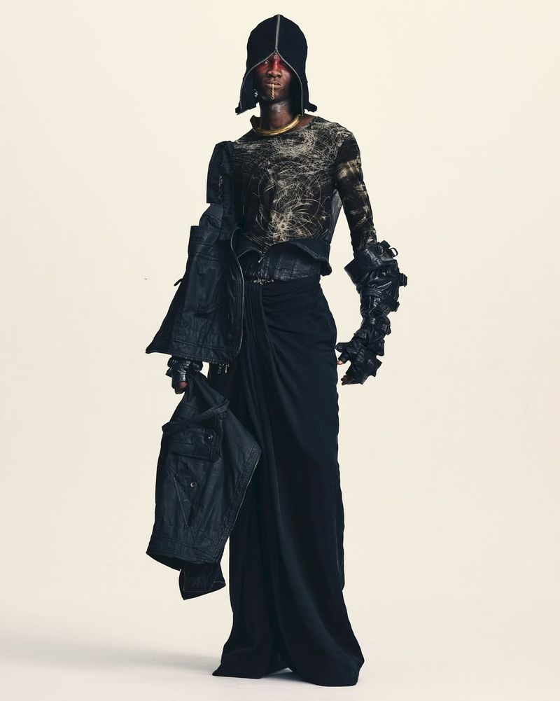 Model wearing an avant-garde black hooded look with draped trousers and textured longsleeve against a plain light studio backdrop.