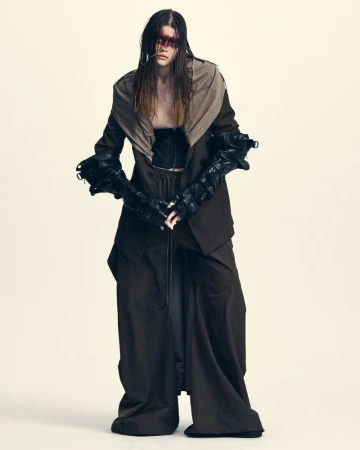 Model in an oversized dark suit with draped beige collar and black leather gauntlet gloves against a pale studio background.