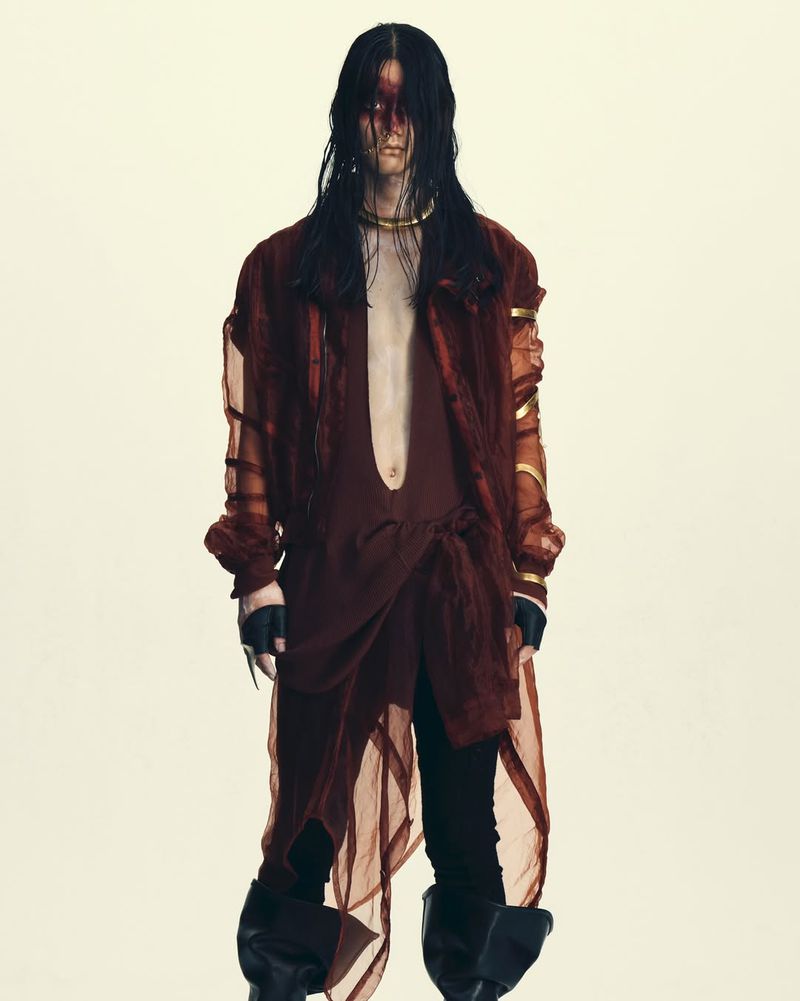 Model with long dark hair wearing a sheer burgundy jacket, open tunic, dark trousers, and tall boots on light background