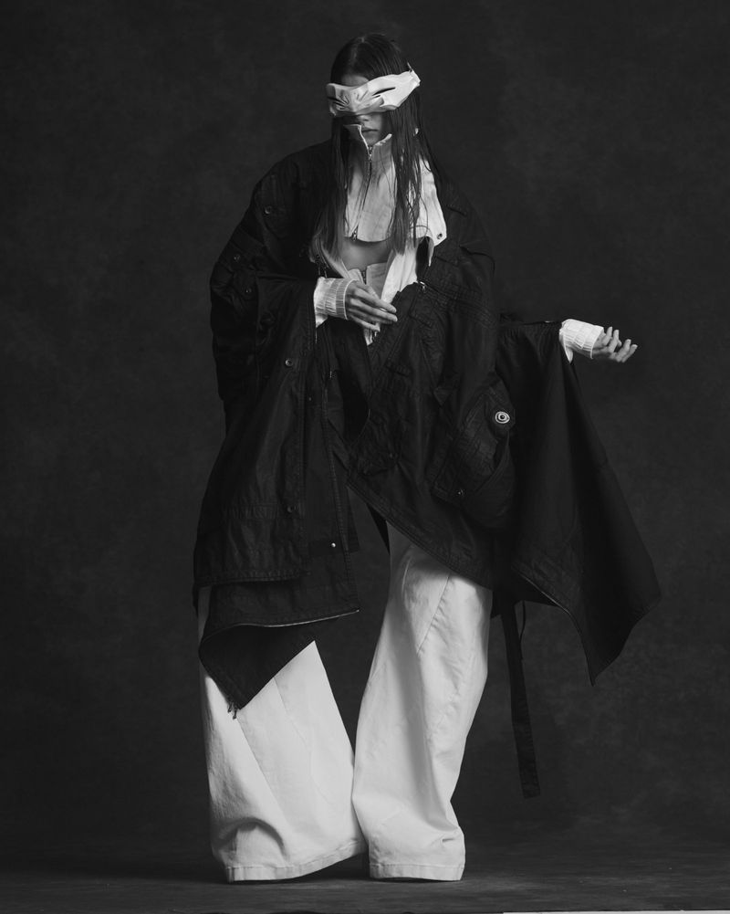 Model wearing a mask and layered black and white clothing against a textured dark background