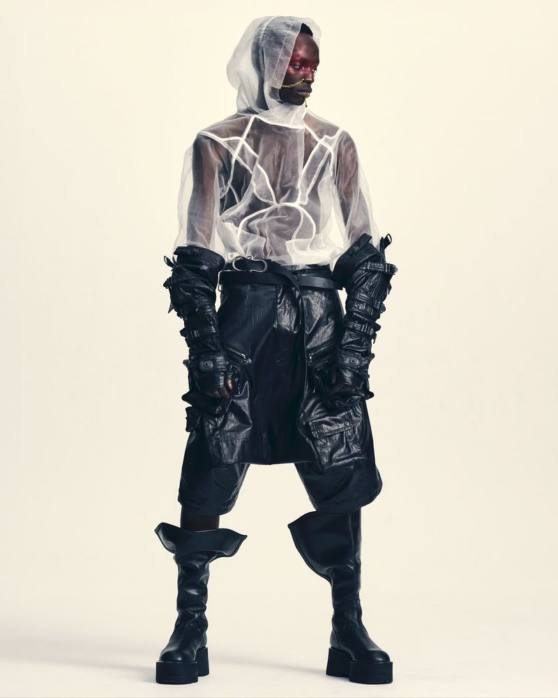 Model wearing a sheer white hooded top and black leather layered garments against a plain white background.