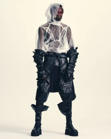 Model wearing a sheer white hooded top and black leather layered garments against a plain white background.