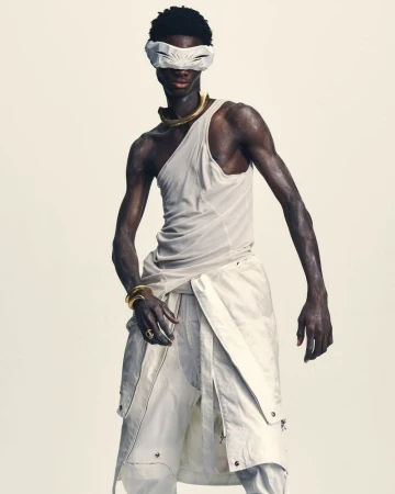 Black male model in an asymmetrical draped tank top, white layered pants, and futuristic white visor against a pale studio backdrop.