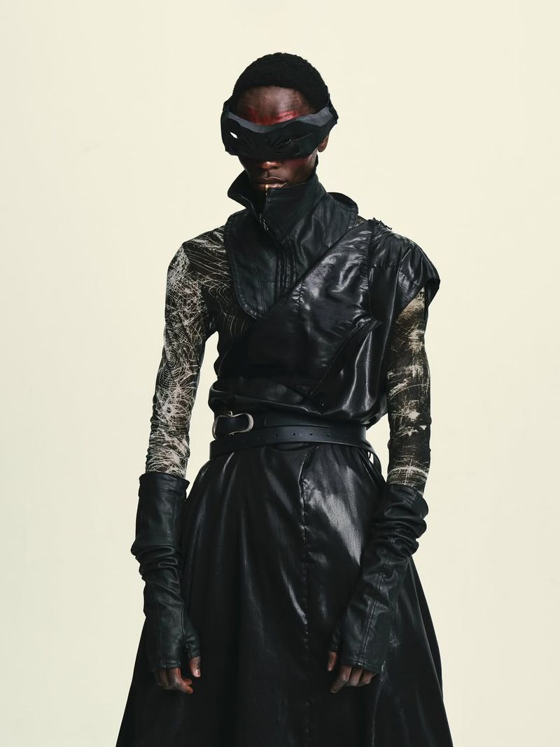 Black male model in an avant-garde black leather outfit and dark futuristic eye mask against a pale studio backdrop.