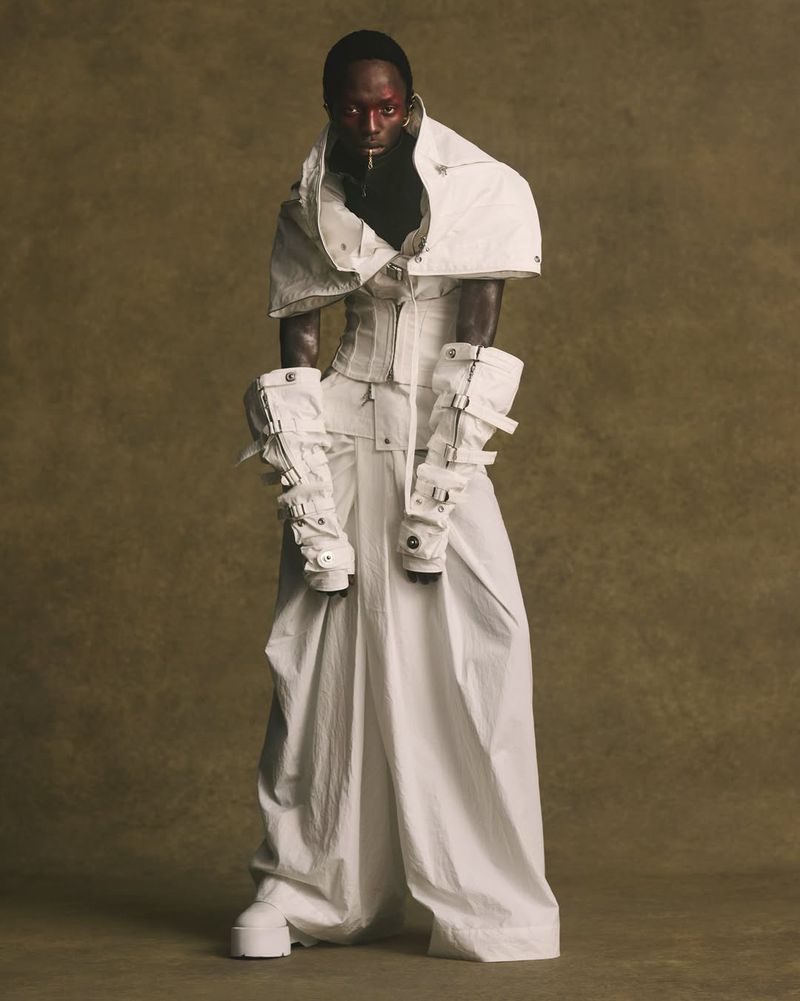 Model wearing an avant-garde white layered sculptural outfit with buckled arm gauntlets against an earthy textured studio backdrop.