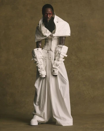 Model wearing an avant-garde white layered sculptural outfit with buckled arm gauntlets against an earthy textured studio backdrop.