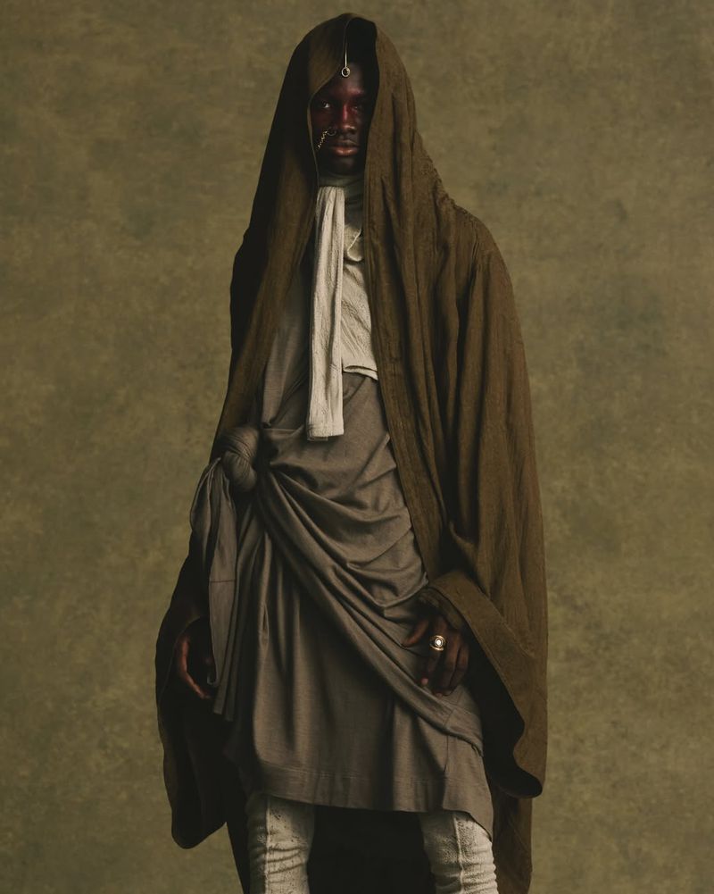Model wearing a draped olive green hooded robe and layered grey tunic against a textured neutral background for JULIUS