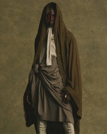 Model wearing a draped olive green hooded robe and layered grey tunic against a textured neutral background for JULIUS