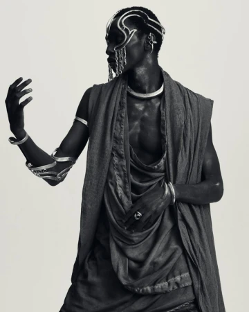 Black male model in draped sleeveless tunic wearing sculptural silver face jewelry and arm cuffs against a light studio backdrop.