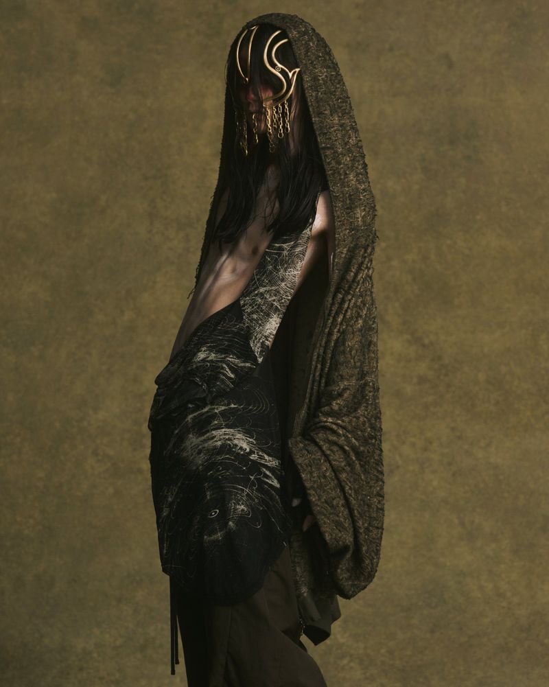 Model wearing a hooded cloak and gold face ornament for JULIUS Spring/Summer 2026