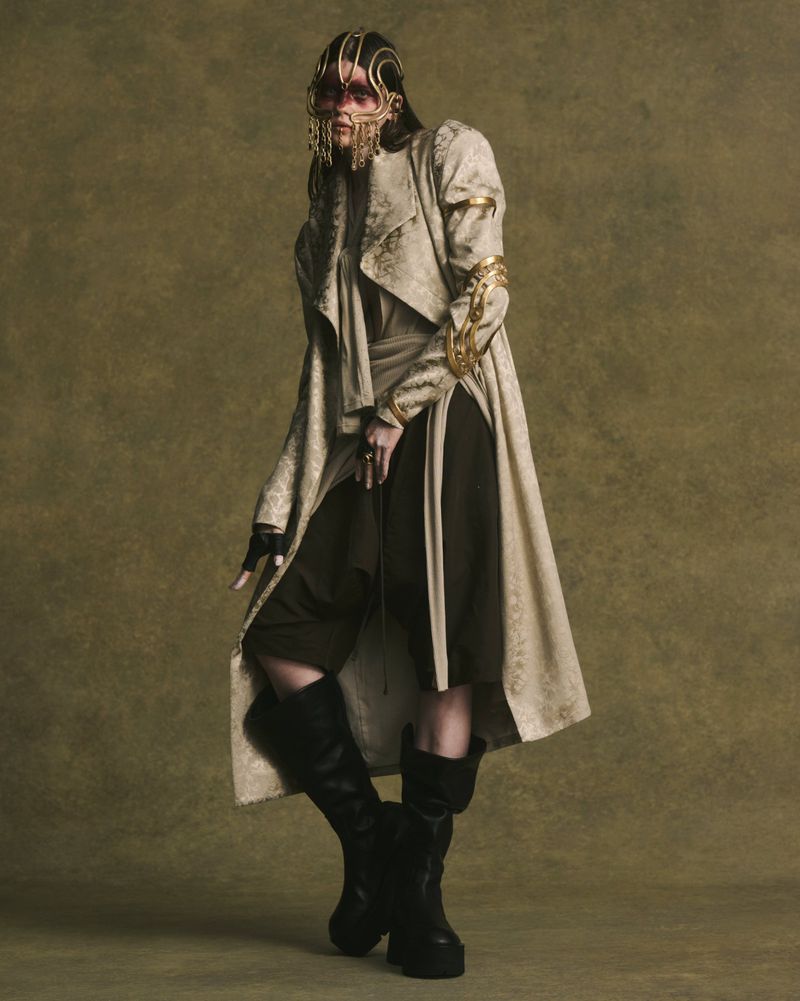 Model wearing patterned coat and gold face ornament with dark boots against olive textured backdrop