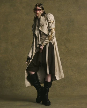 Model wearing patterned coat and gold face ornament with dark boots against olive textured backdrop