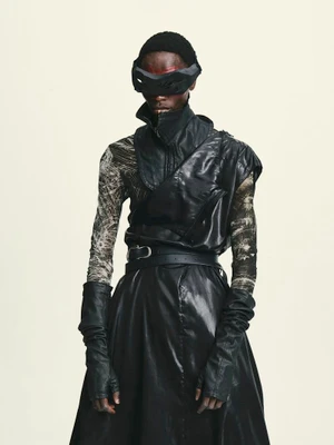 Black male model in an avant-garde black leather outfit and dark futuristic eye mask against a pale studio backdrop.