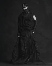 Model wearing a draped all-black avant-garde outfit with long gloves and dark face paint against a textured studio backdrop.